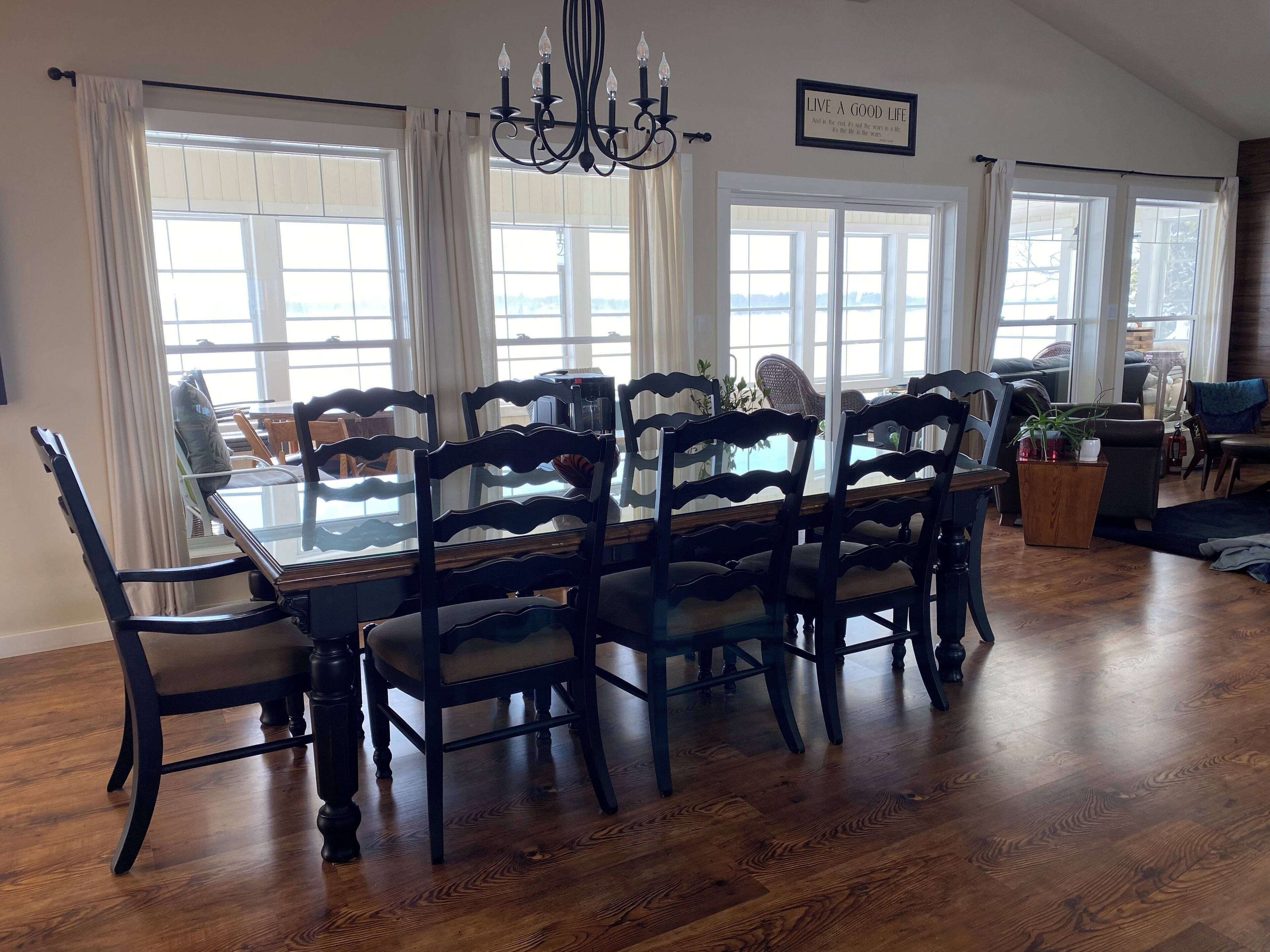 A dining area with a glass-top table surrounded by chairs, large windows with curtains, a chandelier, and a sign that says "LIVE A GOOD LIFE."
