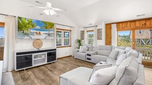 Ein geräumiges Wohnzimmer mit einem großen grauen Sofa, einem Deckenventilator und einem großen Fenster mit Blick auf Grünflächen.