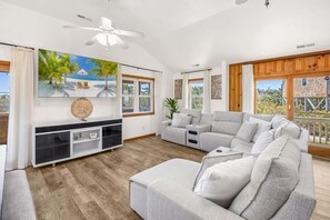 A spacious living room with a large grey sofa, a ceiling fan, and a large window with a view of greenery.