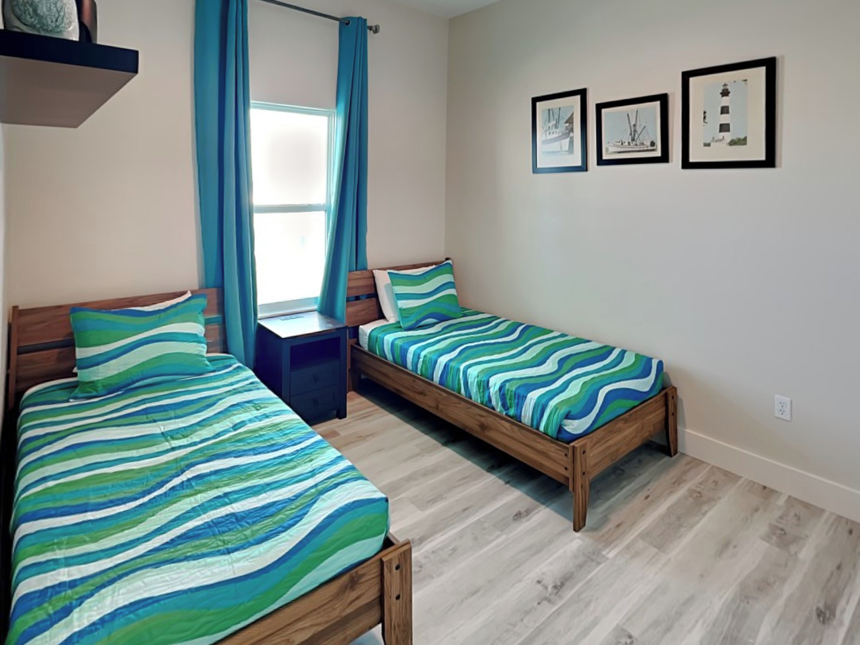 A room with two wooden beds, each with striped bedding, a small table, and a window with curtains. There are three framed pictures on the wall.