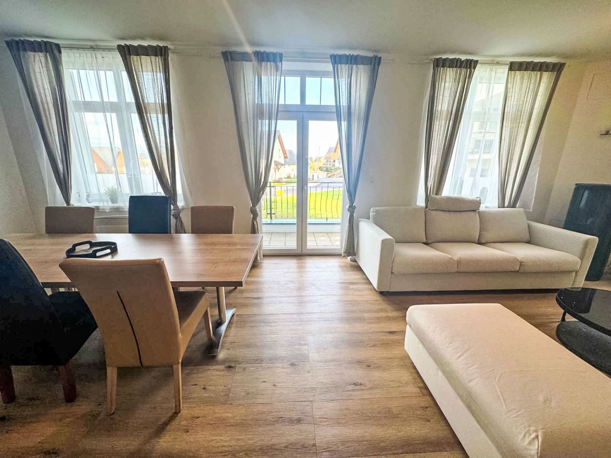 Una sala de estar con piso de madera, un sofá color beige, una mesa de comedor con sillas y ventanas grandes con cortinas.
