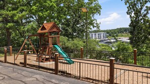 Une aire de jeux avec un toboggan et une structure d’escalade, entourée d’une clôture et d’arbres.