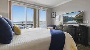 A bedroom with a large bed, a flat-screen TV, a dresser, and a view of a lake through a sliding door. - Land's End #732 Gorgeous Remodel! Parking Level! (Osage Beach)