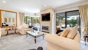A spacious living room with a beige sofa, a coffee table, a TV, and a fireplace. - Orewa Beachside Escape | Backyard & BBQ (Auckland Hibiscus Coast)