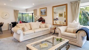 A spacious living room with a beige sofa set, a glass coffee table, and a large mirror. - Orewa Beachside Escape | Backyard & BBQ (Auckland Hibiscus Coast)