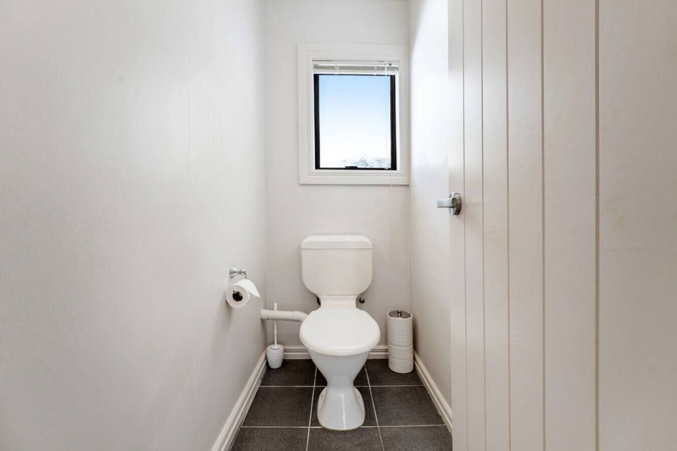 A small, clean bathroom with a toilet, a toilet paper holder, a window, and a trash can.