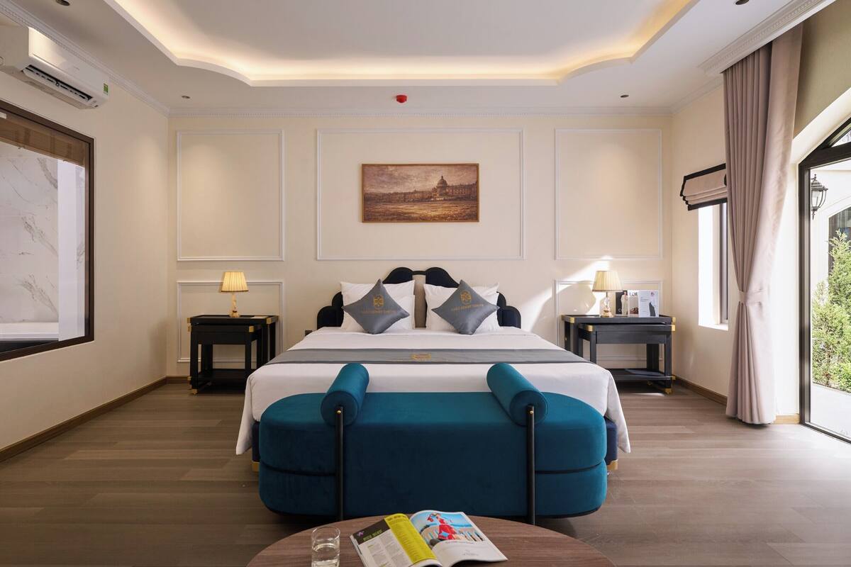 A hotel room with a large bed, a desk, a chair, a table with a magazine and glass, and a painting on the wall.
