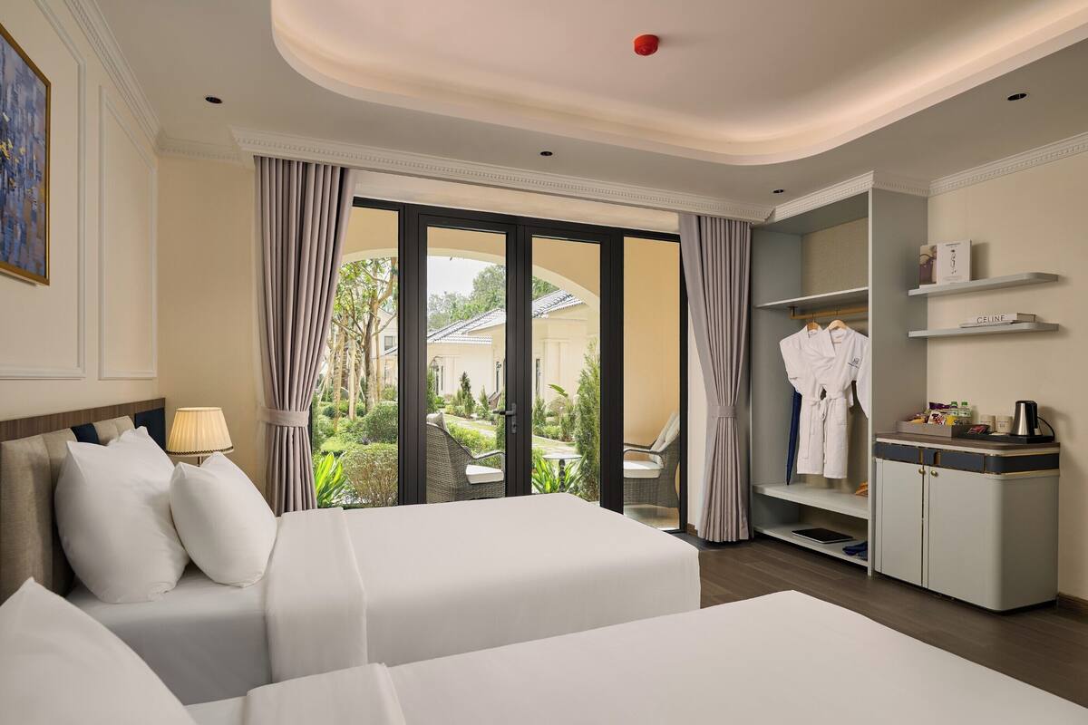 A hotel room with two beds, a wardrobe, a desk with toiletries, and a view of the garden.