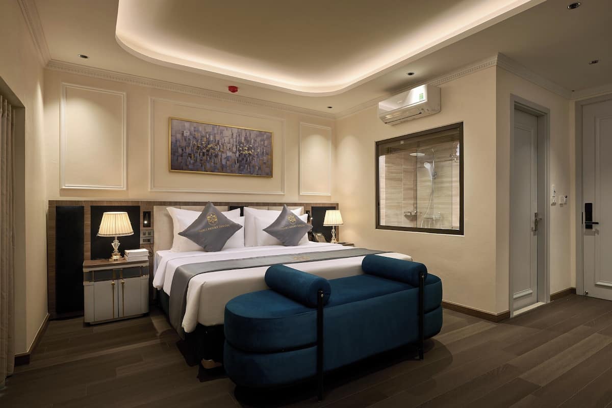 A hotel room with a large bed, a bedside table with a lamp, a blue sofa, a painting on the wall, and a shower area visible through a window.