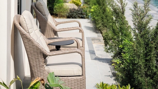 A row of wicker lounge chairs with cushions by a poolside, a clear view of the pool, and a row of houses in the background.