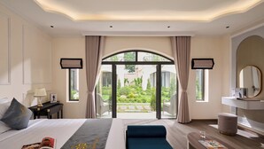A hotel room with a bed, a reading area, a view of the garden, and a bathroom with a mirror.