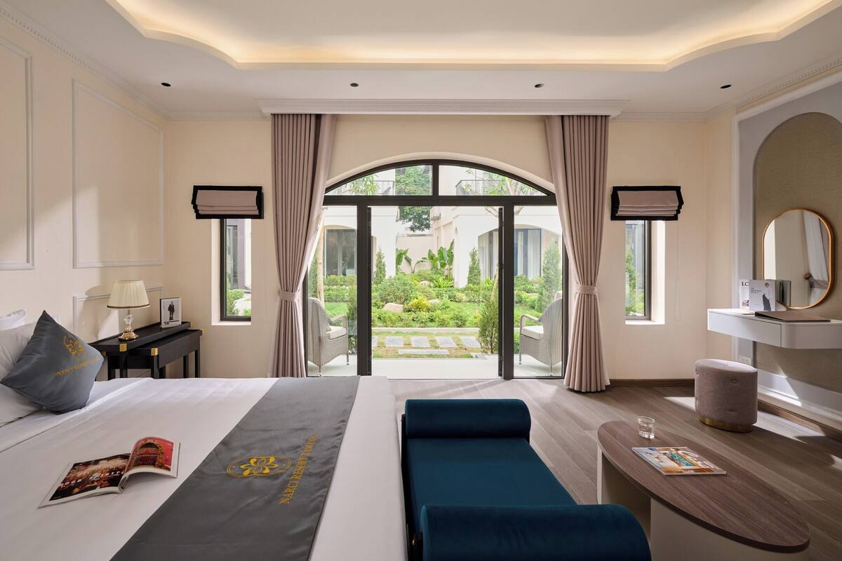 A hotel room with a bed, a reading area, a view of the garden, and a bathroom with a mirror.
