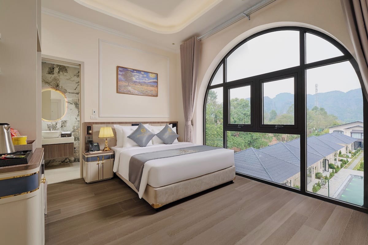 A hotel room with a large bed, a nightstand with a lamp, a bathroom with a marble wall, and a view of a residential area through a large window.