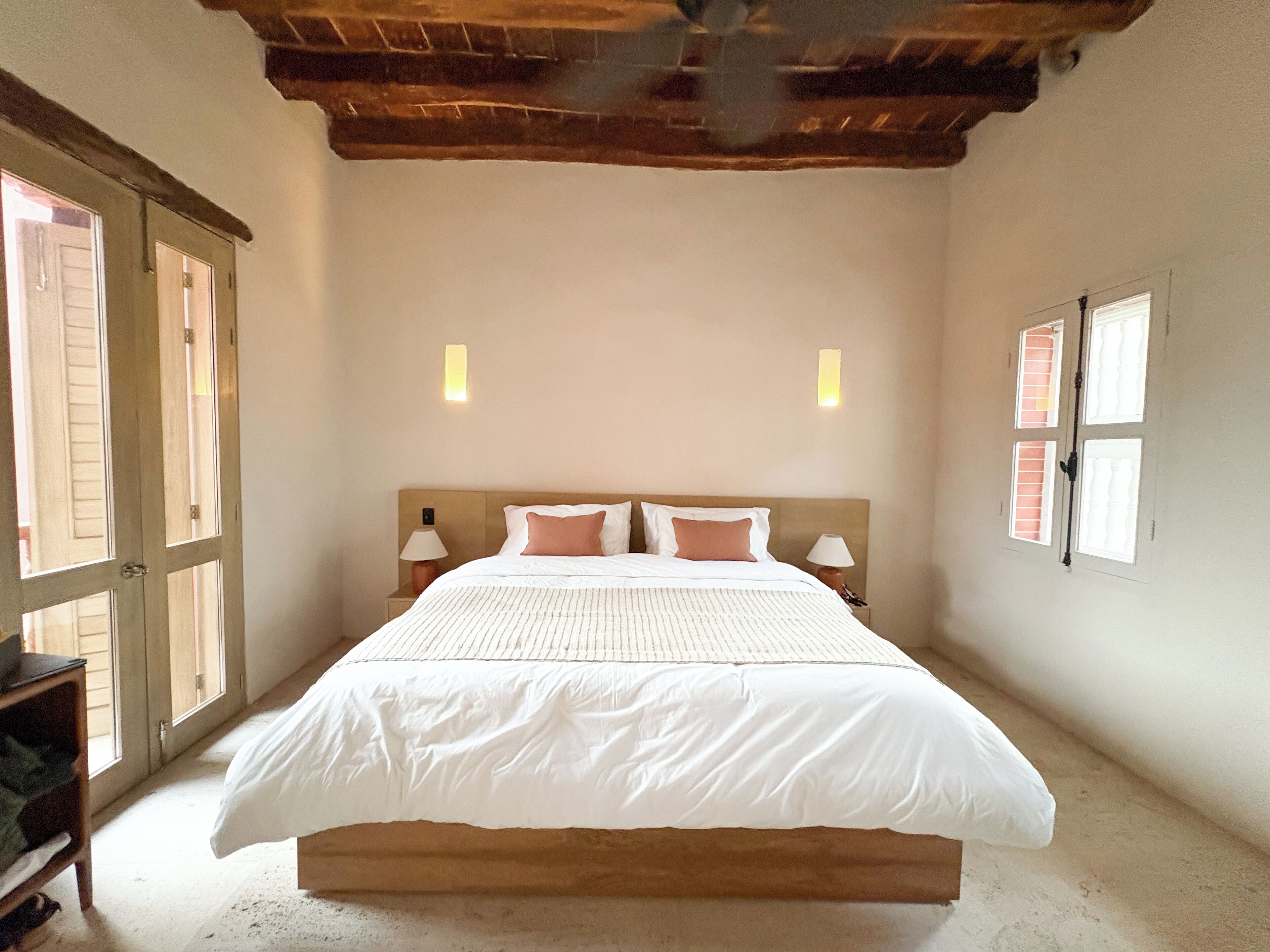 Una camera da letto con soffitto in legno, un letto grande con biancheria bianca, due abat-jour e una finestra con persiane.
