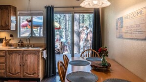 A cozy kitchen with wooden cabinets, a dining area with a table and chairs, and a view of a forest through large windows. - Cozy 2-BR/2-BA cabin in Big Bear perfect for a relaxing, close to the slopes (Big Bear Lake)