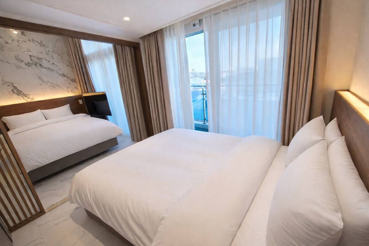 Habitación de hotel con una cama grande, una mesita de noche, un cabecero y vistas al exterior a través de cortinas translúcidas.