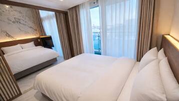 Habitación de hotel con una cama grande, una mesita de noche, un cabecero y vistas al exterior a través de cortinas translúcidas.