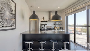 Una cucina moderna con isola centrale, sgabelli da bar, illuminazione a sospensione e un'ampia finestra vista strada.