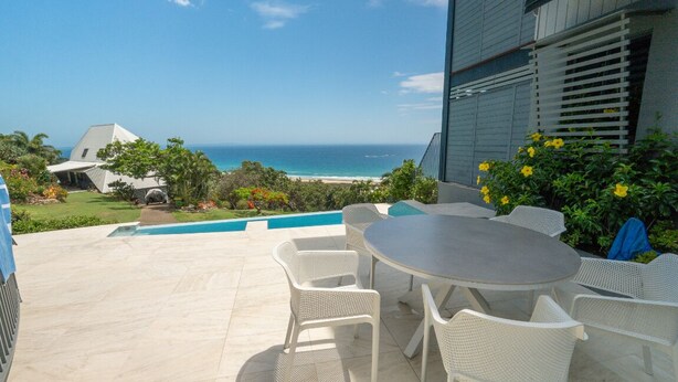 Un patio con una mesa redonda y sillas de mimbre blanco, con vista a una playa y el océano.