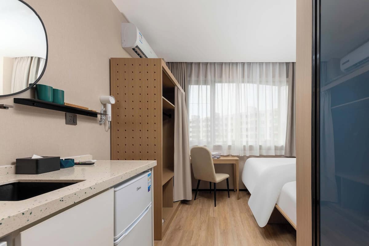 Een hotelkamer met een kitchenette, een bed, een bureau en een raam met dunne gordijnen.