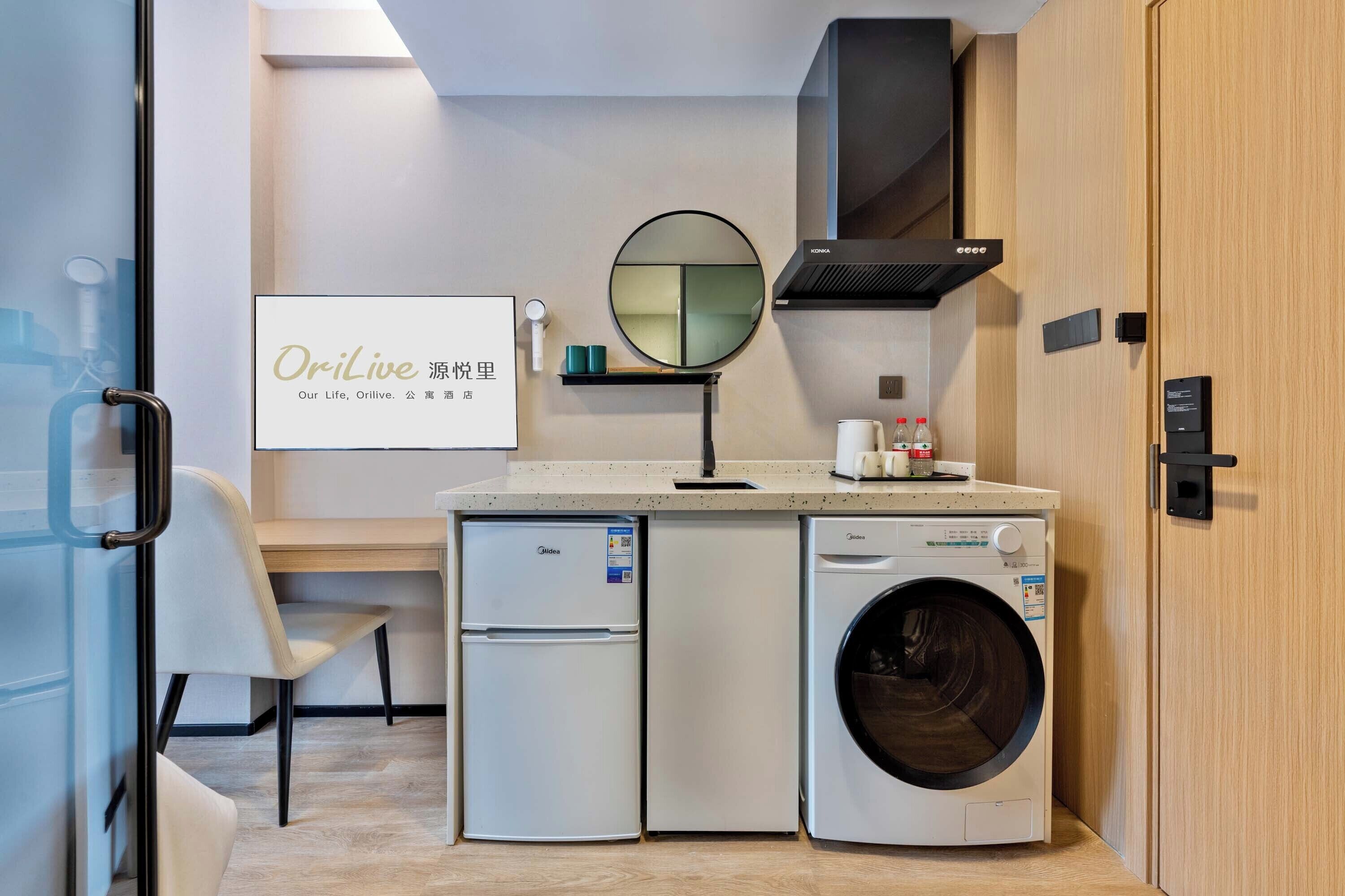 A compact kitchen with a refrigerator, washing machine, and a small countertop with a mirror and a shelf.