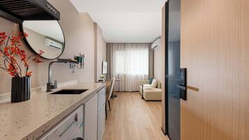 Habitación de hotel pequeña con kitchenette, lavadora, lavamanos, espejo, ventana con cortinas y puerta.