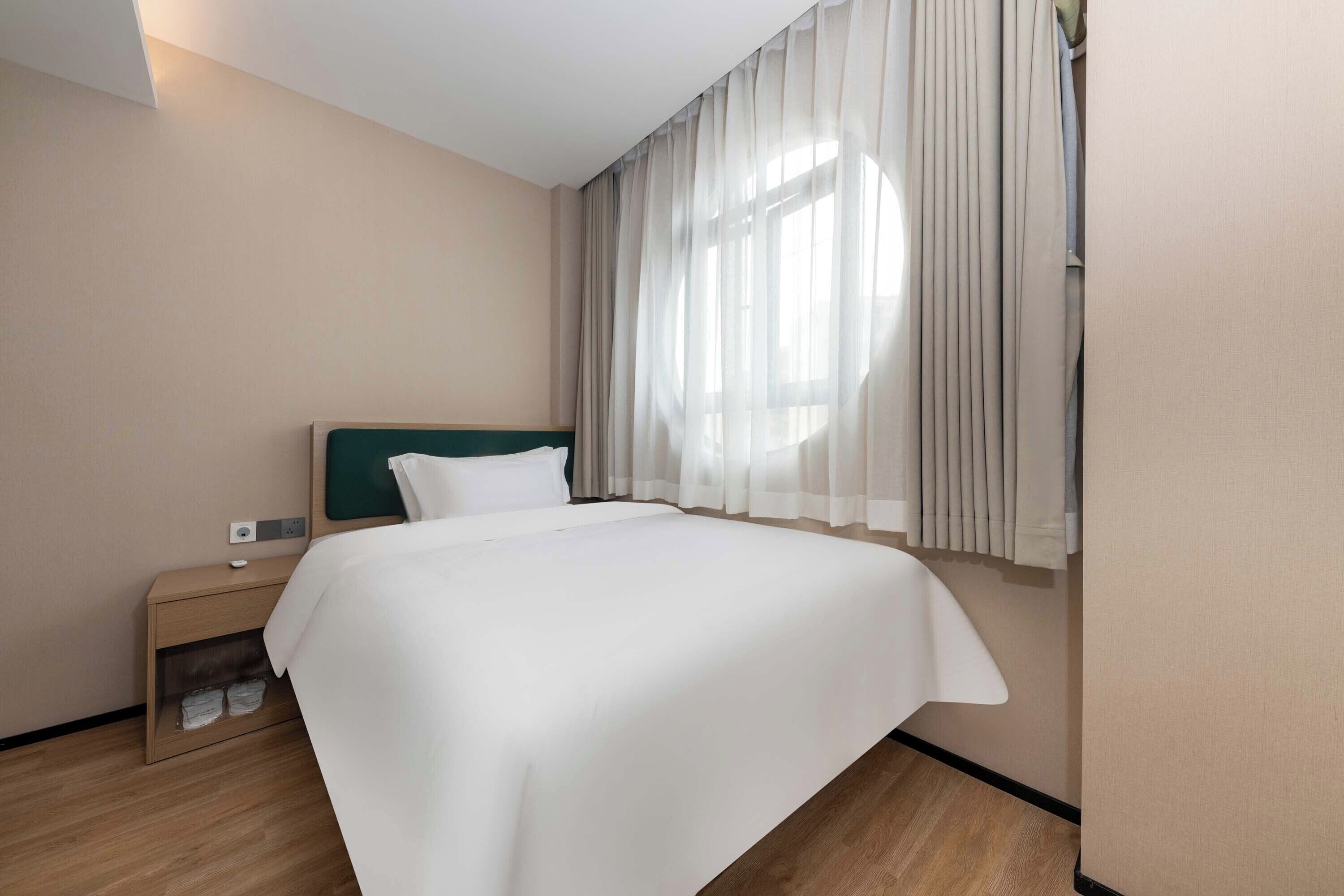 A hotel room with a large bed, a bedside table, a nightstand, and a window with sheer curtains.