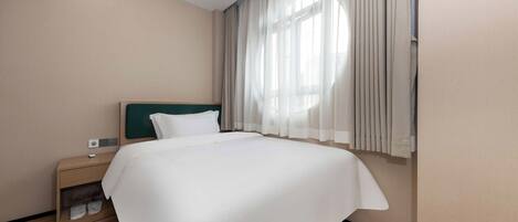 A hotel room with a large bed, a bedside table, a nightstand, and a window with sheer curtains.