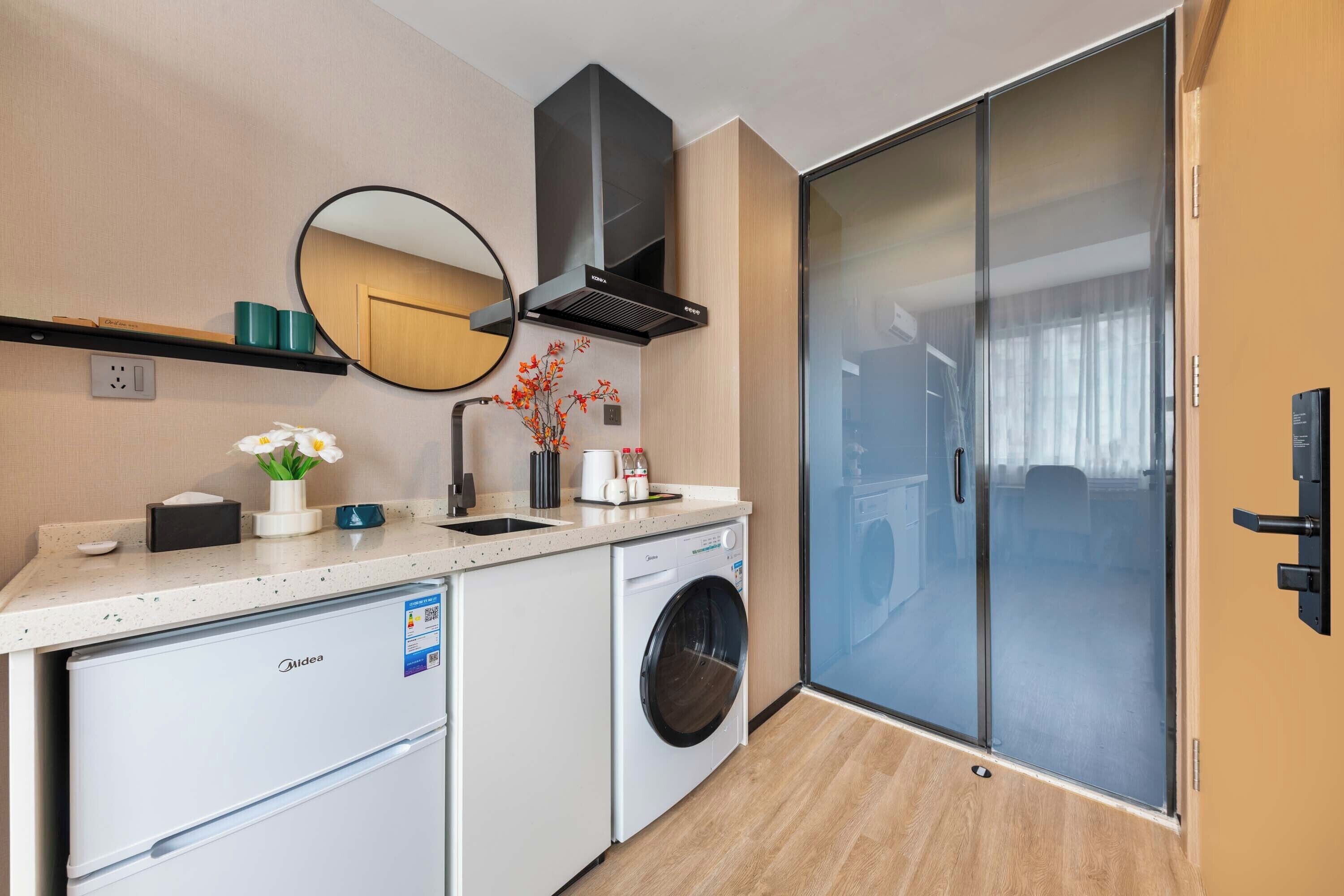 A compact kitchen with a washing machine, a microwave, and a small refrigerator. There is a mirror above the sink and a sliding glass door leading to a balcony.