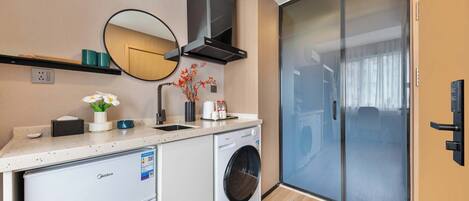 A compact kitchen with a washing machine, a microwave, and a small refrigerator. There is a mirror above the sink and a sliding glass door leading to a balcony.