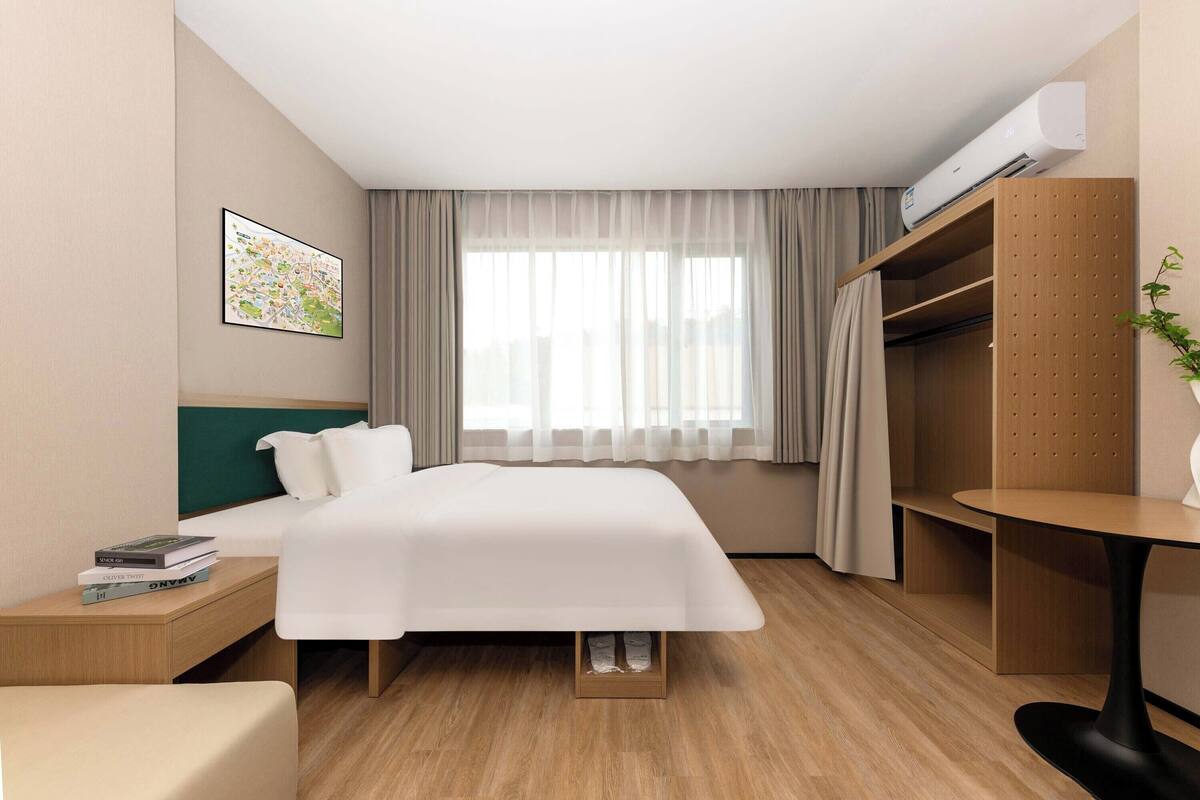 Een hotelkamer met een bed, een nachtkastje met boeken, een bureau met stoel, een kledingkast en een raam met gordijnen.
