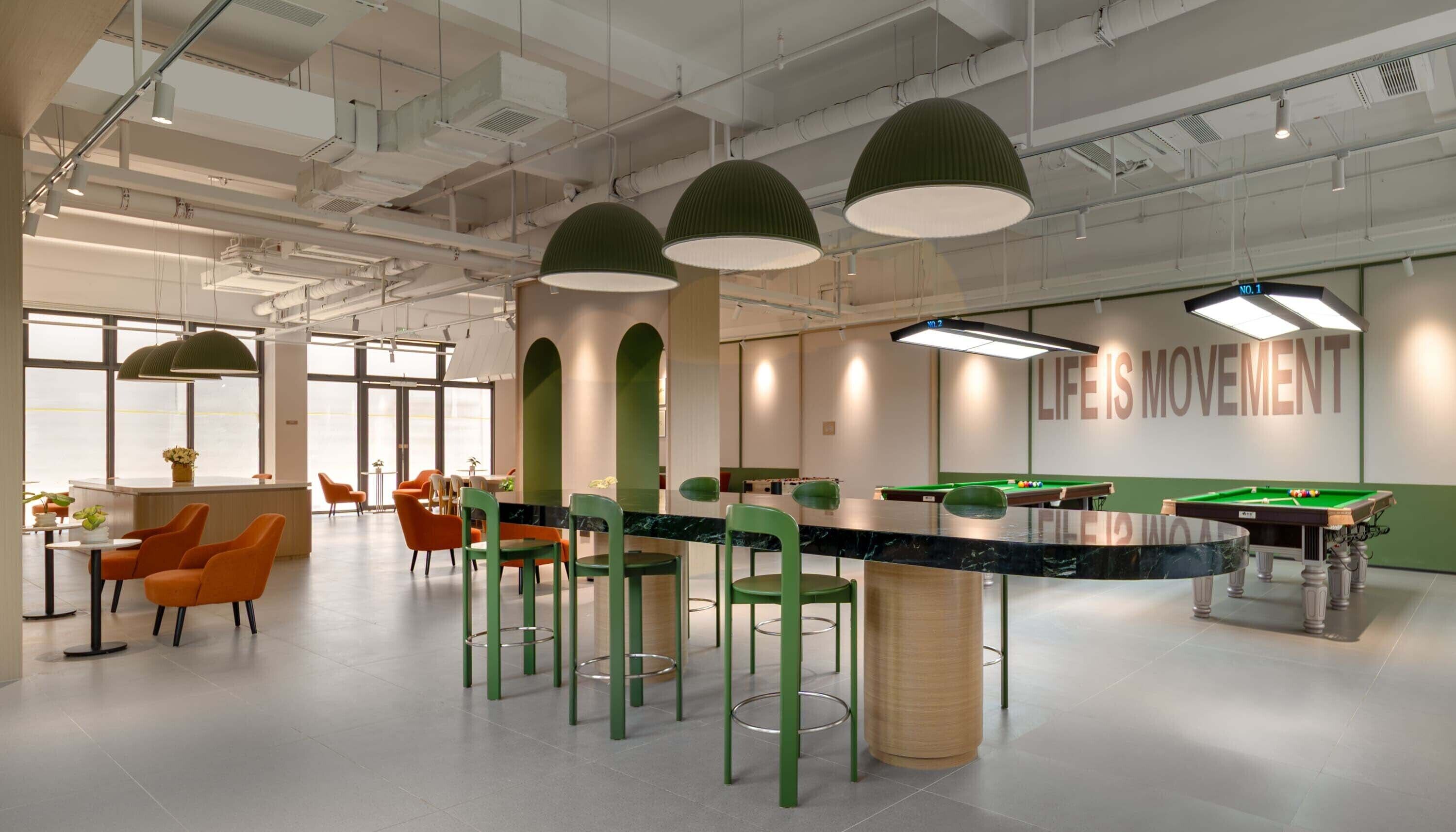 A modern indoor space with a pool table, green chairs, and a bar area.