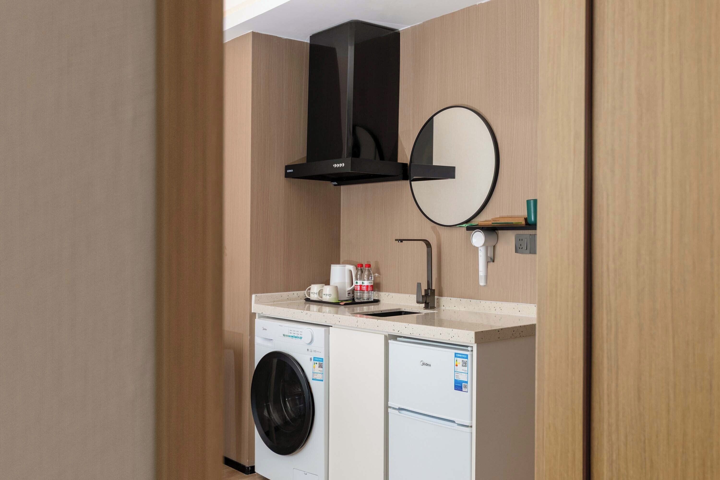 A compact hotel room with a washing machine, a small refrigerator, a sink with a mirror, and a black range hood.
