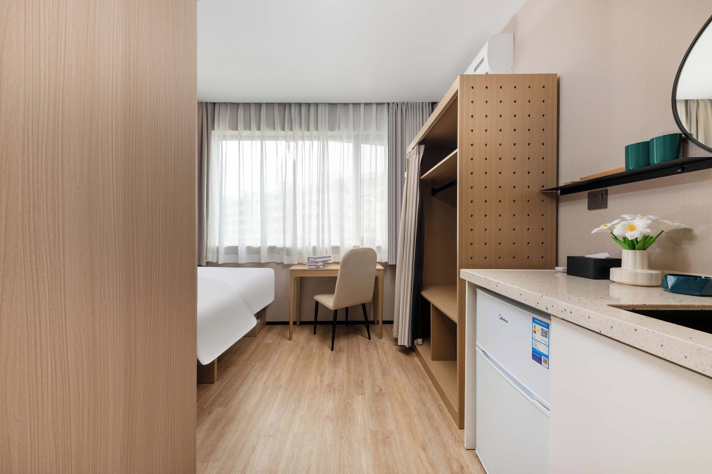 A compact hotel room with a bed, a desk, a chair, a small kitchenette, and a window with sheer curtains.