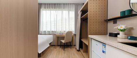 A compact hotel room with a bed, a desk, a chair, a small kitchenette, and a window with sheer curtains.