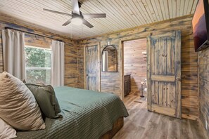 A bedroom with a bed, a ceiling fan, a window with curtains, and wooden walls. - Rustic Retreat — Easy Access, Peaceful Setting, Close to Everything (Sevierville)