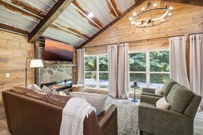 A cozy living room with a fireplace, comfortable seating, and large windows offering a view of the outdoors. - Rustic Retreat — Easy Access, Peaceful Setting, Close to Everything (Sevierville)