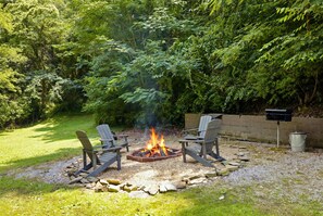Ein Feuerkorb mit zwei Adirondack-Stühlen, umgeben von einem Kiesbereich und einer Rasenfläche, mit dichtem Grün im Hintergrund.