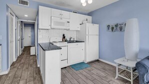 Eine kleine Küche mit weißen Schränken, einer Mikrowelle und einem Kühlschrank. Auf dem Boden liegt eine blaue Matte und es gibt einen kleinen Tisch mit einem Stuhl.