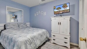 Ein Schlafzimmer mit Bett, Kommode, Spiegel und Fernseher. Auf der Wand steht das Wort 'BEACH'.