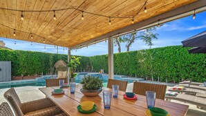 An outdoor patio with a wooden table, wicker chairs, and a view of a swimming pool.