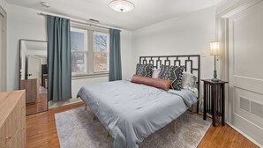 Ein Schlafzimmer mit Bett, Nachttisch, Spiegel, Fenster mit Vorhängen und Deckenleuchte.