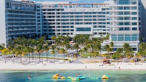 Un hôtel situé en bord de mer, avec plusieurs étages, des palmiers et une mer d’un bleu limpide.