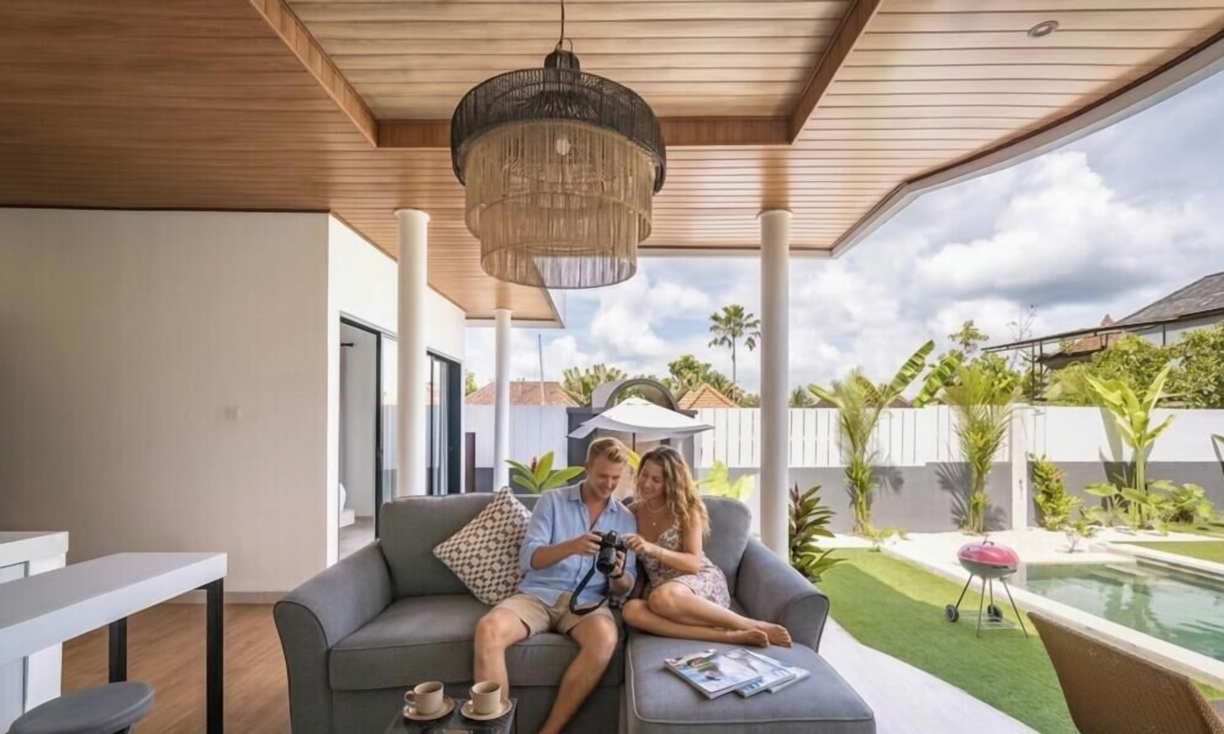 Et moderne udendørs opholdsområde med træloft, hængende lampe og udsigt til swimmingpool og grøn bevoksning.