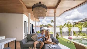 Et moderne udendørs opholdsområde med træloft, hængende lampe og udsigt til swimmingpool og grøn bevoksning.