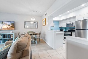 A modern kitchen with white cabinetry, stainless steel appliances, and a dining area with a wooden table and chairs. - Sandpiper Towers 408 (Satellite Beach)