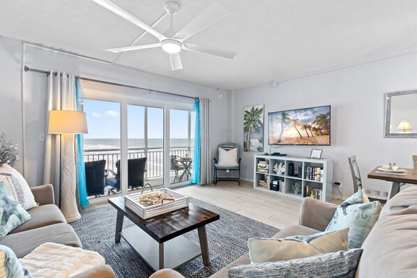 A spacious living room with a large window overlooking the beach, a ceiling fan, a TV, and a bookshelf. - Sandpiper Towers 408 (Satellite Beach)