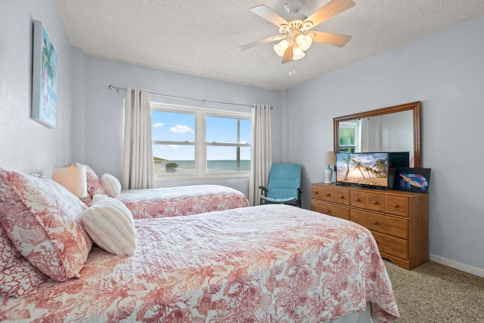 Une chambre avec deux lits, un ventilateur de plafond, une commode, une chaise et une grande fenêtre donnant sur l’océan.