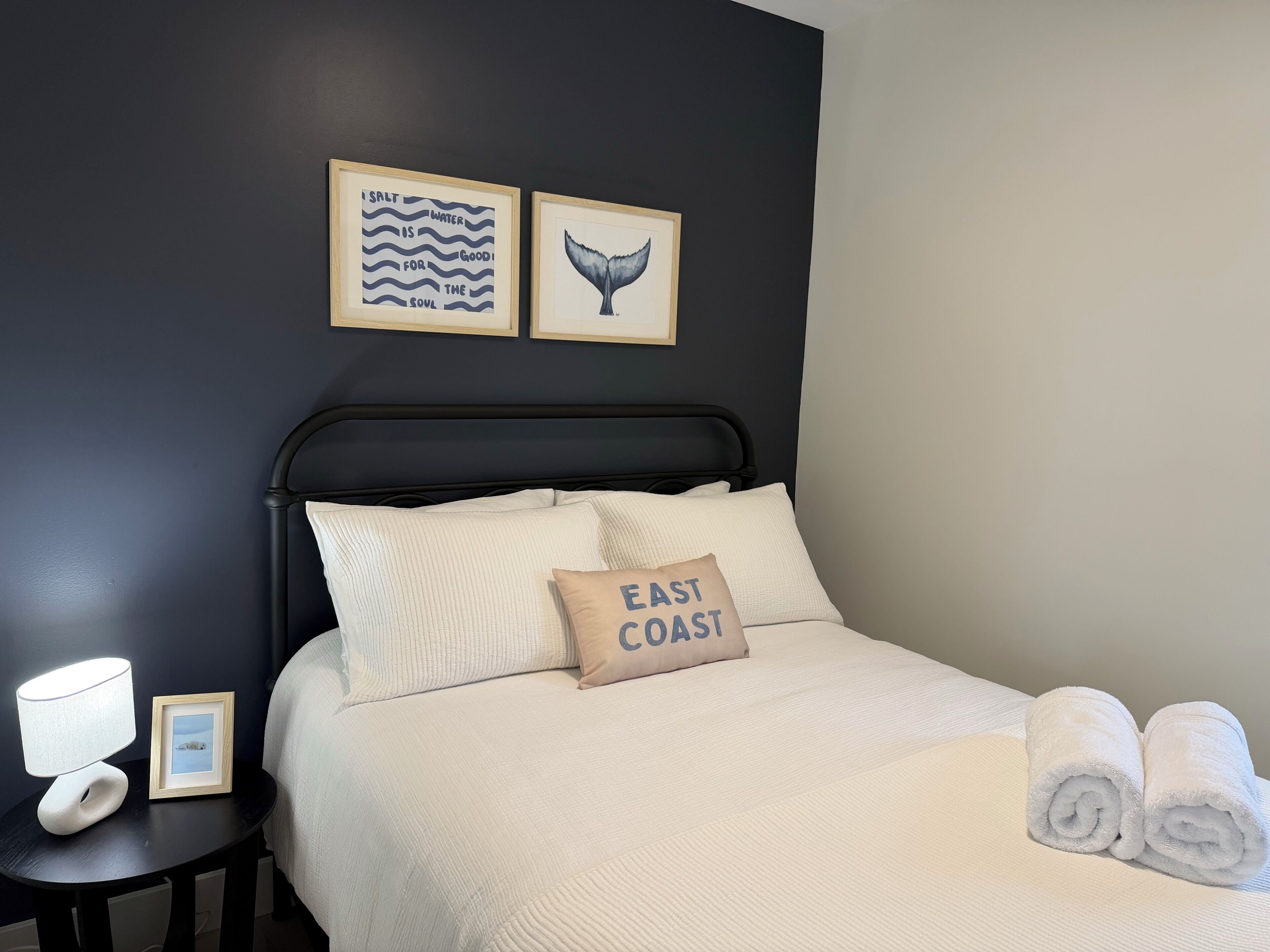 Ein ordentlich bezogenes Bett mit einem Kissen mit der Aufschrift 'EAST COAST', einem Bilderrahmen mit dem Schwanzflossenbild eines Wals und einem gerahmten Zitat über Salz und Wasser an der Wand.