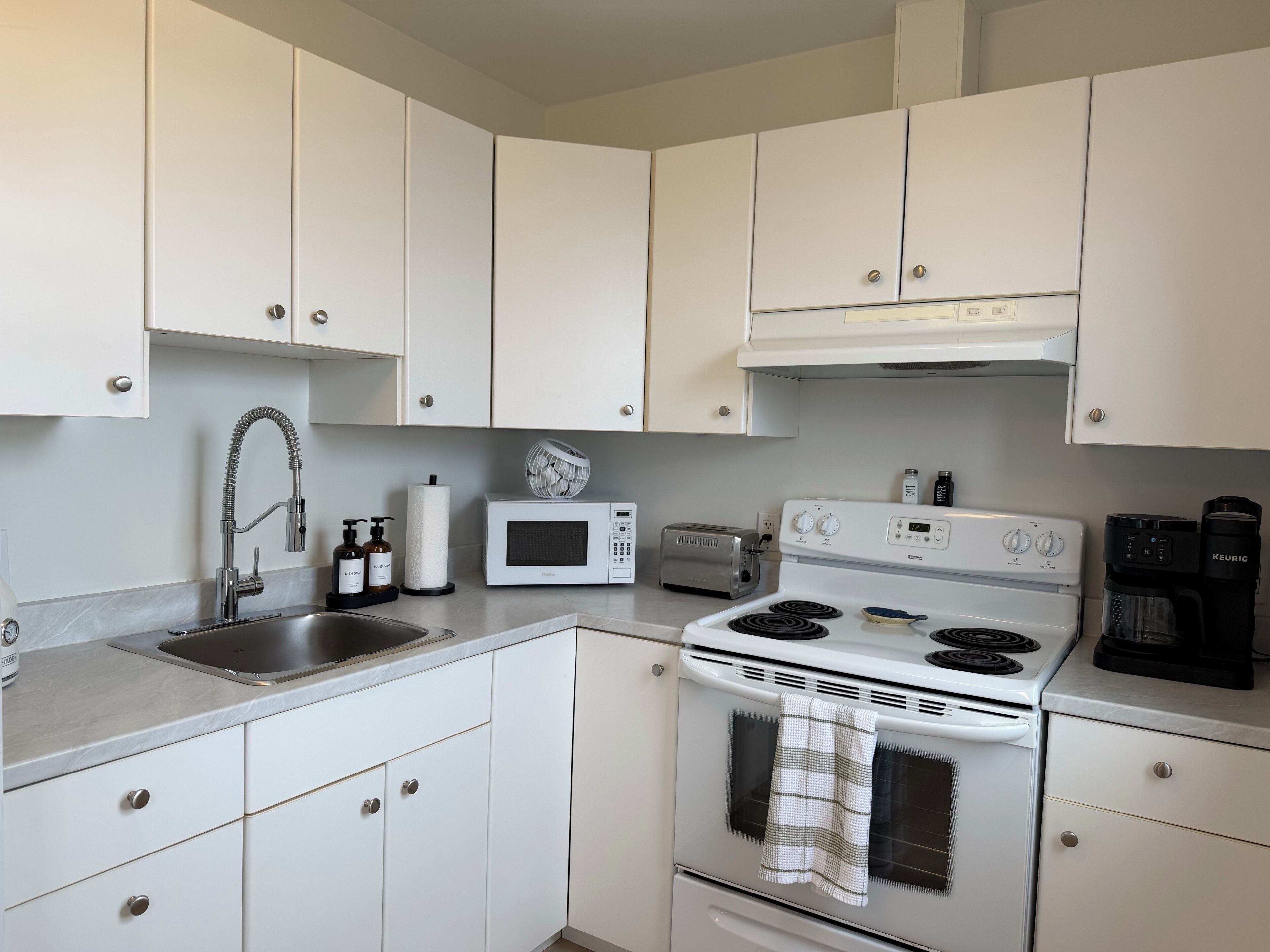 Une cuisine compacte avec des meubles blancs, un évier en acier inoxydable, un four à micro-ondes, un grille-pain, une cuisinière et une machine à café Keurig.